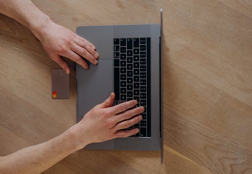 A person using a laptop and credit card for online shopping, viewed from above on a wooden table.